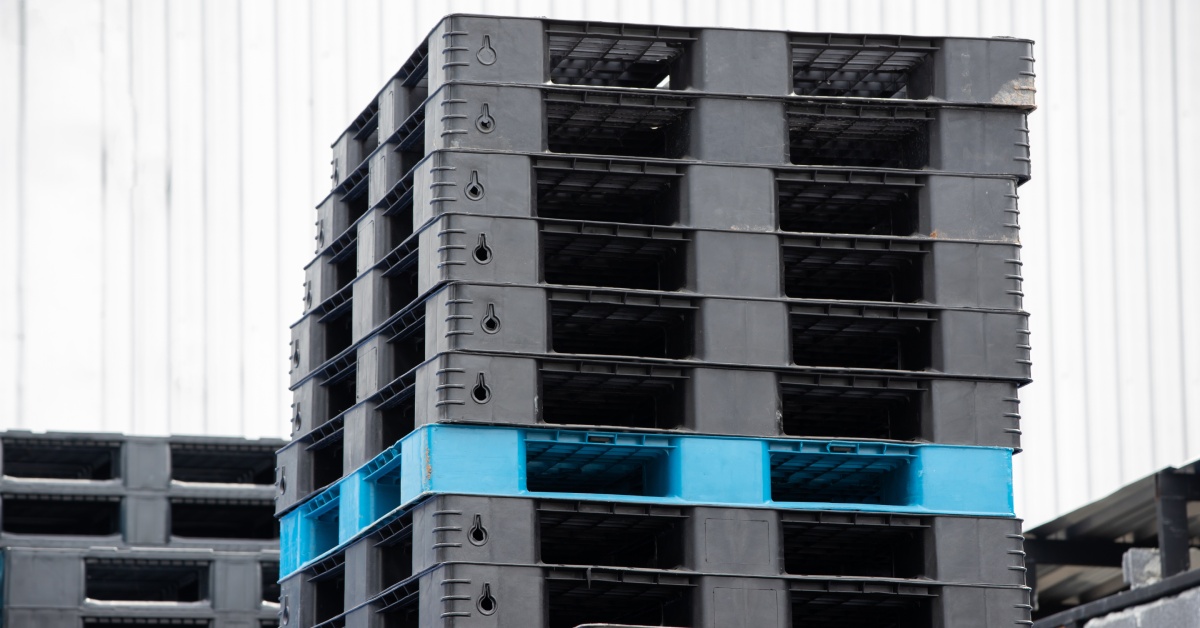 A warehouse contains a group of black plastic pallets and one light-blue plastic pallet stacked on top of one another.