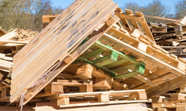 A stack of wooden pallets sitting in a large pile of wood debris outside. Trees and blue sky are visible in the background.