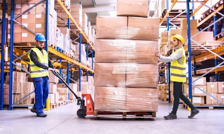 Two warehouse employees, one male and one female, moving a loaded pallet truck across a distribution facility.