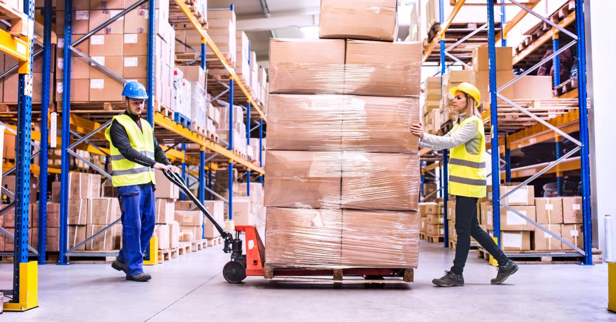 Two warehouse employees, one male and one female, moving a loaded pallet truck across a distribution facility.