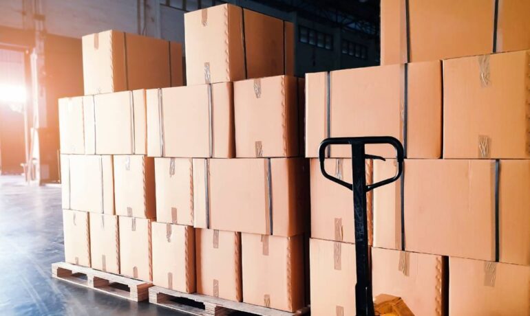 Many shipment boxes sit on top of wooden pallets inside a warehouse storage facility with a hand pallet truck on the right side.