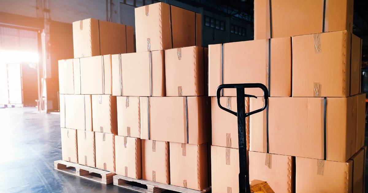 Many shipment boxes sit on top of wooden pallets inside a warehouse storage facility with a hand pallet truck on the right side.