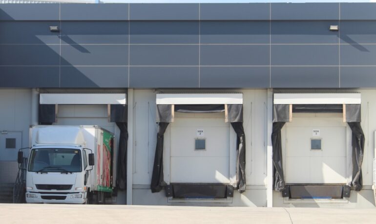 A building with blue panels on top has three loading docks. A truck is exiting the dock on the left.