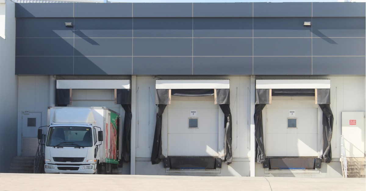 A building with blue panels on top has three loading docks. A truck is exiting the dock on the left.