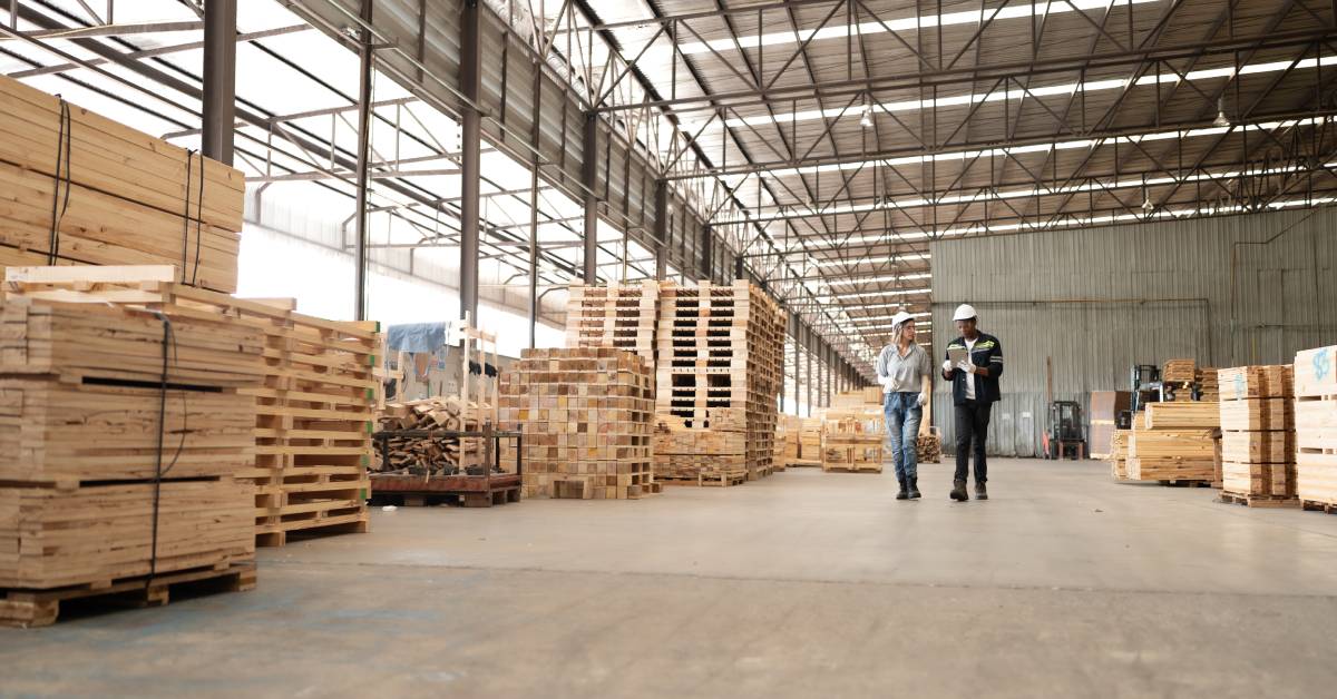 Two workers wearing white hard hats walk through a warehouse containing large quantities of wooden pallets.