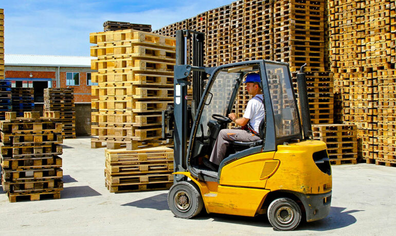 A man wearing a blue hard hat drives a yellow forklift. Large stacks of wood pallets are to his left and right.