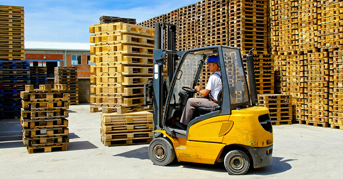 A man wearing a blue hard hat drives a yellow forklift. Large stacks of wood pallets are to his left and right.