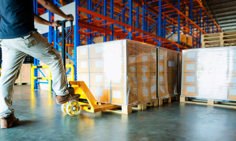 A warehouse worker slides a hand pallet truck under a cargo shipment pallet. Other pallet shipments are on the right.