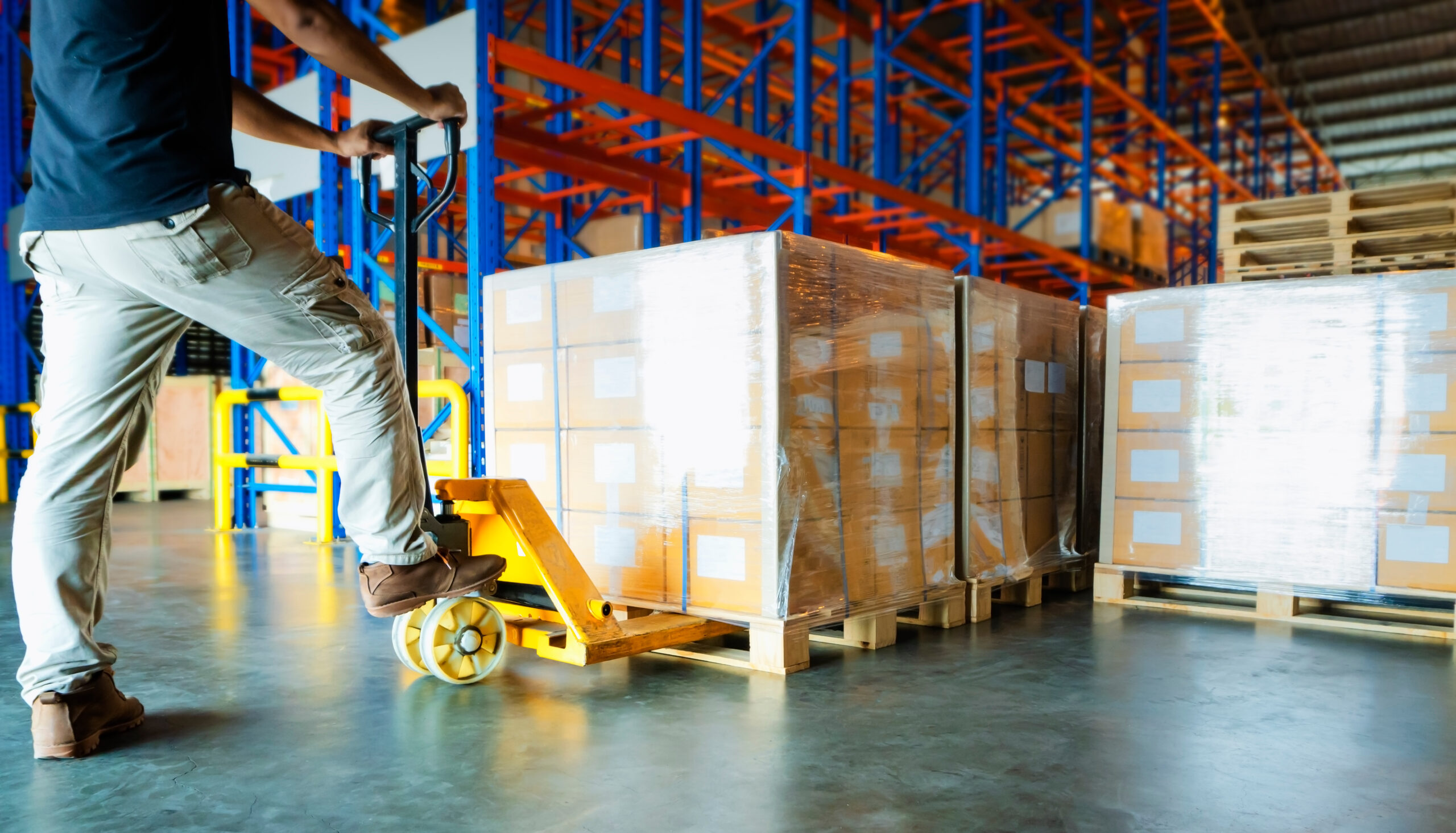 A warehouse worker slides a hand pallet truck under a cargo shipment pallet. Other pallet shipments are on the right.