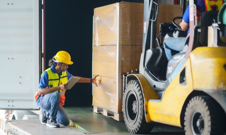 A forklift driver loads a pallet of boxes of goods, while a supervisor inspects the shipment and uses a digital tablet.