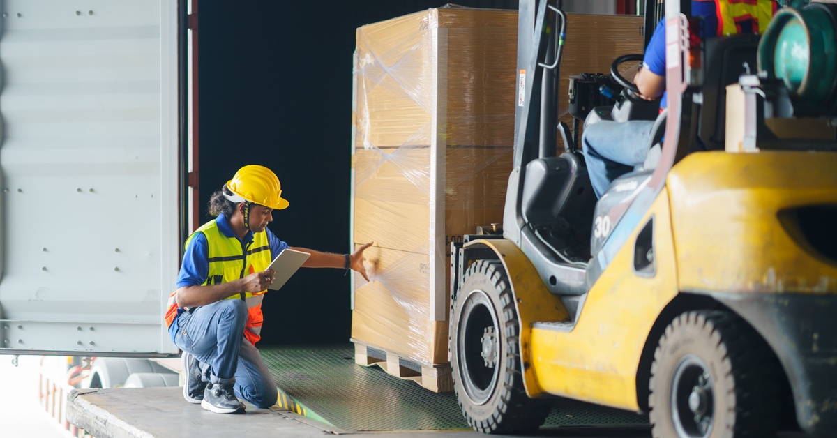 A forklift driver loads a pallet of boxes of goods, while a supervisor inspects the shipment and uses a digital tablet.